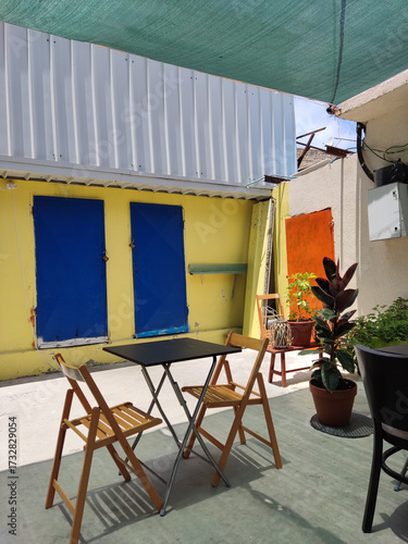 A vibrant and eclectic outdoor patio scene featuring a small black metal bistro table with two light wooden folding chairs, set against a bold, colorful architectural backdrop 