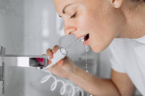 A young woman uses a water flosser to clean her teeth in the bathroom in the morning. Concept: morning routine and oral hygiene at home.