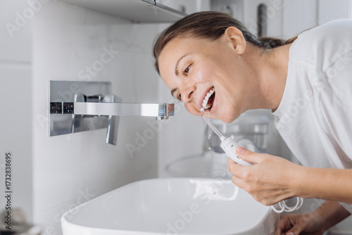 A young woman uses a water flosser to clean her teeth in the bathroom in the morning. Concept: morning routine and oral hygiene at home.