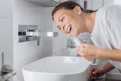 A young woman uses a water flosser to clean her teeth in the bathroom in the morning. Concept: morning routine and oral hygiene at home.