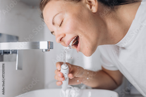 A young woman uses a portable water flosser for oral care in the bathroom. Concept: personal hygiene and modern dental care routine.