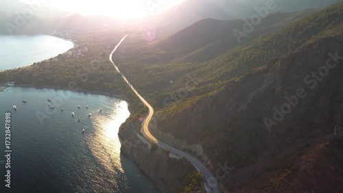 An aerial view captures the scenic coastal road winding along the Datça Peninsula, leading towards the historic ruins of Knidos Ancient City in Turkey. The golden hour light illuminates the winding pa