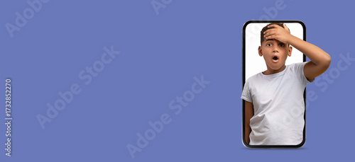 Photos A young boy looks shocked and surprised as he holds his forehead, appearing to react to something on his smartphone