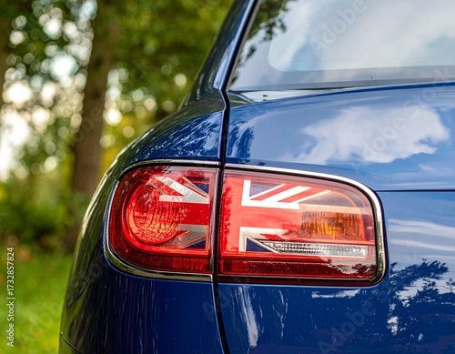 Close-up of the rear side of a glossy blue vehicle with chrome-framed tail light and Union Jack emblem. Orange and red light sections. British design, automotive detail, outdoor lighting.