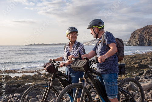 Senior active couple riding on the pebble beach with electric bicycles at sunset. Authentic elderly retired life and sustainable mobility concept