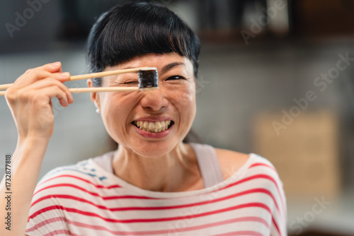 Cheerful woman holding sushi roll with chopsticks in front of her eye, smiling playfully at home in the kitchen. Fun portrait of Asian woman enjoying Japanese food.
