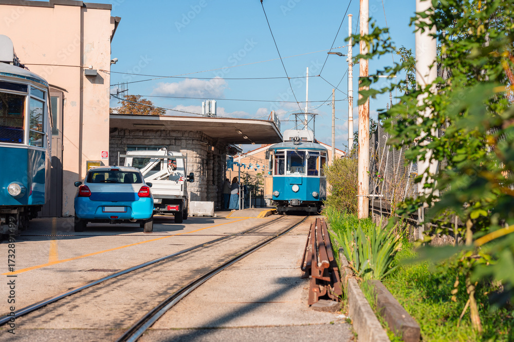 Naklejka premium Historic blue Opicina tram arriving at a small station in Opicina, Italy. The vintage streetcar passes along narrow tracks near parked cars, with sunlight casting warm tones on the scene.
