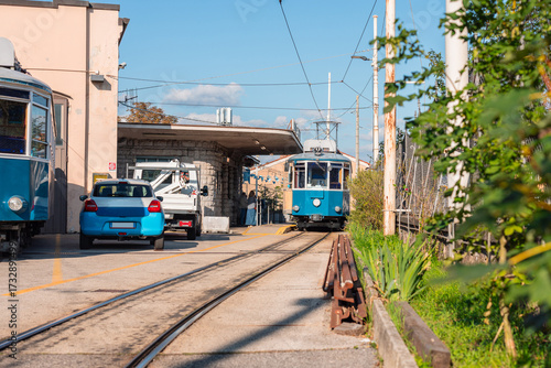 Historic blue Opicina tram arriving at a small station in Opicina, Italy. The vintage streetcar passes along narrow tracks near parked cars, with sunlight casting warm tones on the scene.