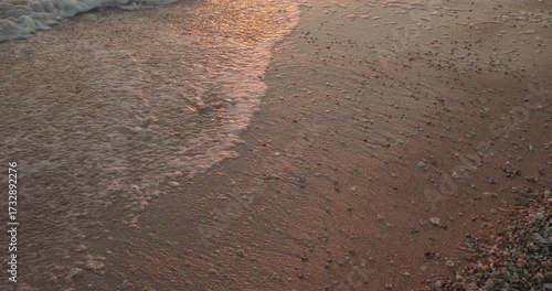Beautiful ocean waves gently washing over a sandy shore with small pebbles, reflecting the warm, golden light of a spectacular sunset, creating a serene and tranquil summer evening mood