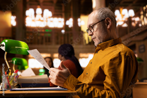 Elderly man with a serious expression studies at a college library desk, engaged in academic research and reading textbooks to complete his degree and enrich his understanding of literature.