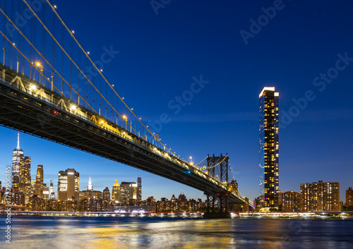 View of New York City and the Brooklyn Bridge with city lights at night near the water, at night.