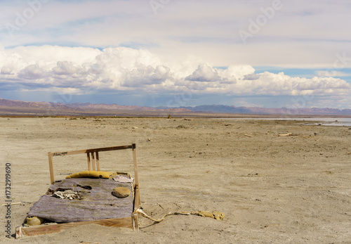 Fotografie A decaying bed sits abandoned in a vast, desolate desert landscape under a cloudy sky