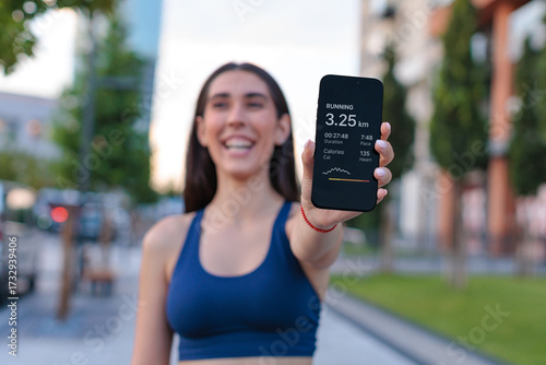 young woman runner smiles and shows distance on smartphone screen during jog in urban park area in summer