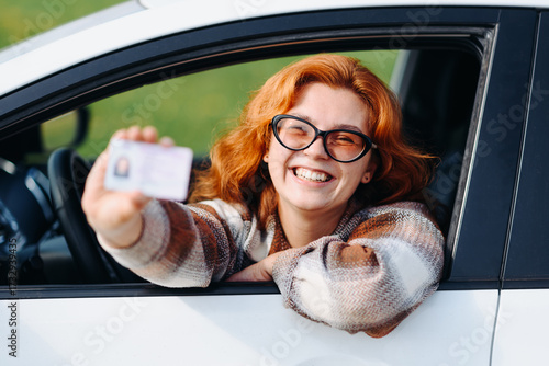 young beautiful redhead caucasian woman shows new driver's license, sitting in white passenger car, beige plaid shirt, leaning out of car window, smiling happily, becoming driver, selective focus