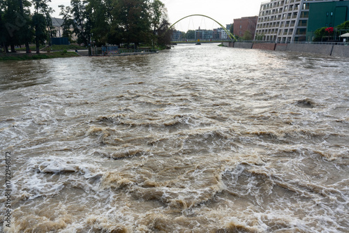 Severe urban river flooding with turbulent muddy brown water submerging park and modern pedestrian bridge with buildings in background. Concept of extreme weather events, urban flood disaster impact.