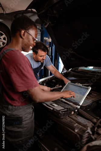 Two mechanics work together on a car engine using a laptop for diagnostics in an auto repair shop
