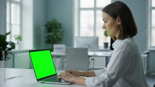 Woman working on a laptop with a green screen in a modern office environment