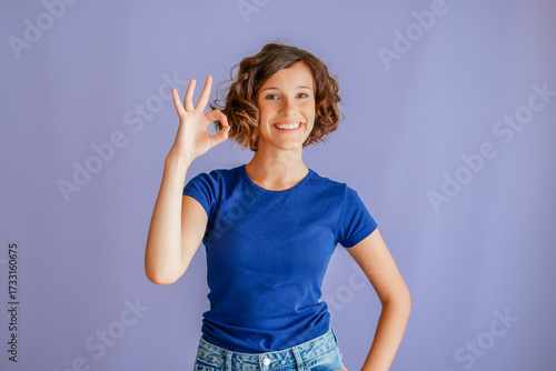Lovely young lady showing OK hand gesture with cheerful expression and positive vibe for approval concepts on vibrant blue studio background with friendly mood