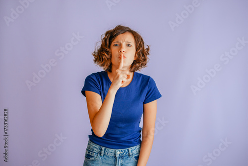 Serious young girl showing secrecy with finger to lips and focused vibe for prohibition on vibrant blue studio background with stern energy