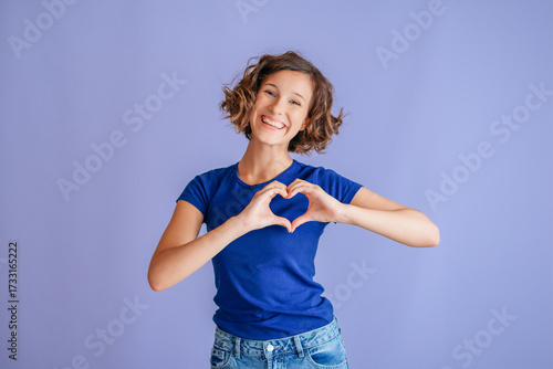 Happy teenage girl making heart gesture with cheerful smile for friendship and support on vibrant blue studio background with joyful expression