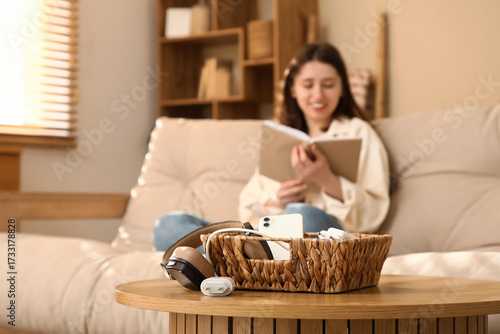 Fotografie Basket with gadgets on table against woman reading book at home, closeup