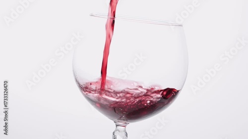 Red wine slowly pouring in glass over white background. Close-up of red wine forms beautiful wave in glass. Grape wine pouring in glass at white background. 4k footage.