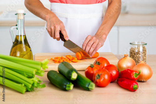 A person preparing a healthy meal by slicing carrots on a wooden cutting board surrounded by fresh vegetables in a bright kitchen setting