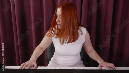 Joyful young redhead woman in elegant white dress playing piano with closed eyes against purple curtain indoors, symbol of inspiration and artistic lifestyle.