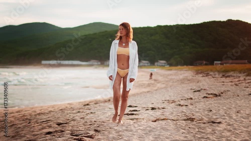 Young woman in yellow bikini and white shirt walks along peaceful beach at sunset, enjoying solitude and connection with nature. Concept of freedom, summer vacation and serene coastal lifestyle.