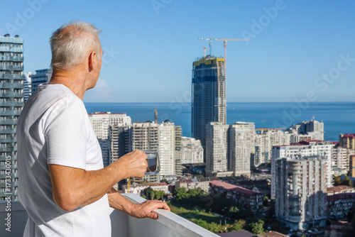 An elderly, confident man, standing on the balcony of a skyscraper with a cup of tea, looks at the view of the coastal city under construction. The concept of real estate investment