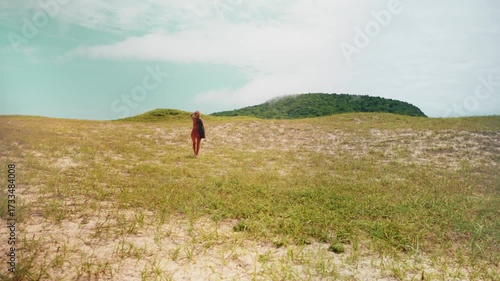 Woman walking alone through grassy dunes with yoga mat on shoulder, embracing peaceful solitude and connection to nature. Symbol of solo travel and emotional balance in natural surroundings.
