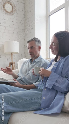 Middle-aged man and woman in casual clothes sitting on sofa with laptop, drinking coffee and discussing at home. Concept of teamwork, communication, planning, lifestyle and collaboration