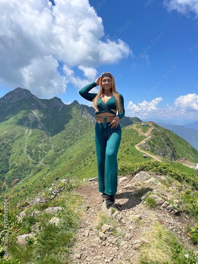 Naklejka premium A beautiful girl in a green suit stands against the backdrop of alpine meadows with flowers in the summer in Krasnaya Polyana. The weather is clear and sunny, with a blue sky.