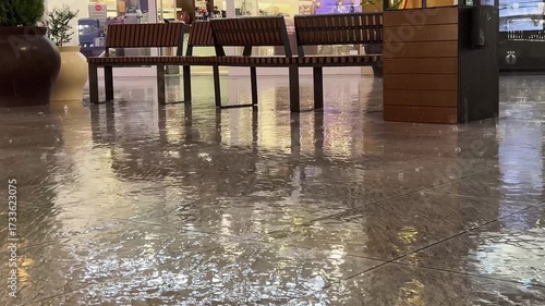 Outdoor benches in a public area, seen on a rainy day. The ground tile reflecting the surrounding benches and lights from nearby shops