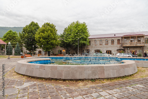 View of a placid fountain reflecting the sky, embraced by stone and verdant trees, with buildings in the backdrop, Goris, Syunik Province, Armenia.