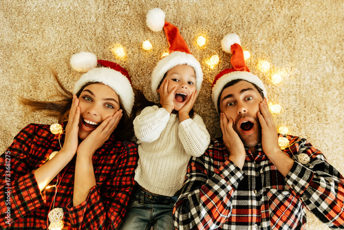 Surprised family lying on the carpet with glowing lights, parents and daughter in Santa hats holding their faces with joy, celebrating love, Christmas eve, and New Year.