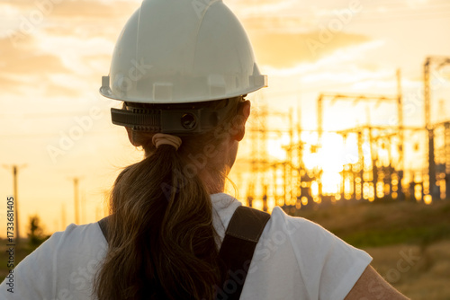 Engineer observes electric grid at sunset while wearing safety gear and a hard hat in a construction site