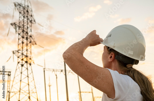 Woman in hard hat observes power lines at sunset near a utility facility while conducting site inspections