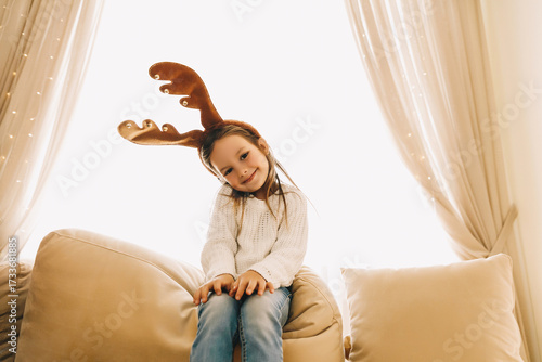 Cheerful little girl in reindeer antlers posing on sofa, dressed in cozy white sweater and jeans. Bright holiday lights and curtains behind create warm festive Christmas mood at home.