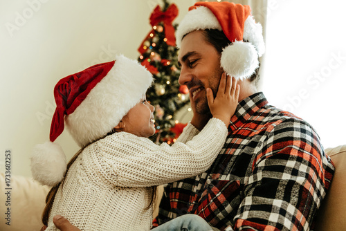 Little girl in Santa hat holding her father’s face with love, both smiling warmly in front of a glowing Christmas tree, sharing a tender holiday moment.
