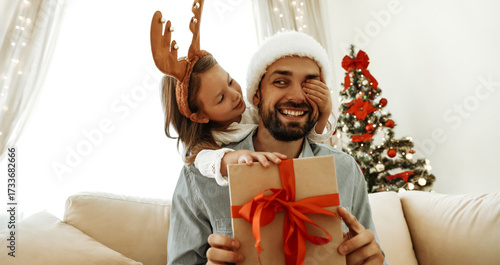 Smiling father in a Santa hat holds a Christmas gift with a red ribbon while his playful daughter in reindeer antlers covers his eye, sharing a joyful holiday moment by the tree.