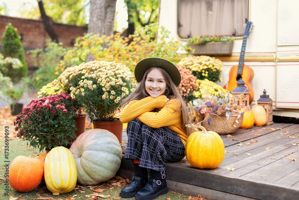 Fototapeta premium Beautiful smile young girl with long hair in knitted clothes with flowers chrysanthemums and pumpkins for Halloween at home. Fall portrait of a pretty teen girl in a cozy orange sweater and green hat 