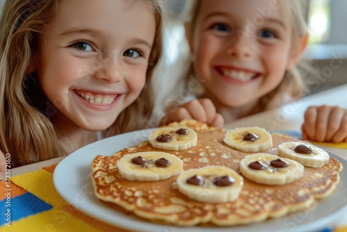 Two happy girls eagerly gaze at delicious pancakes topped with bananas and chocolate, sharing a joyful breakfast moment.