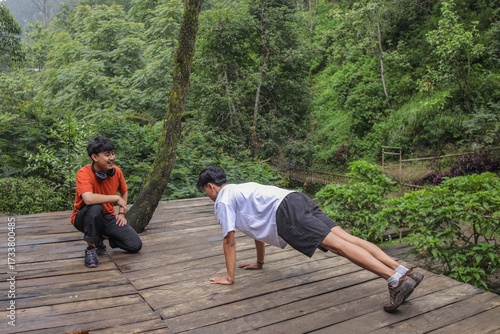 Active Young Man Doing Pushups Outdoors While Friend Watches In Lush Forest