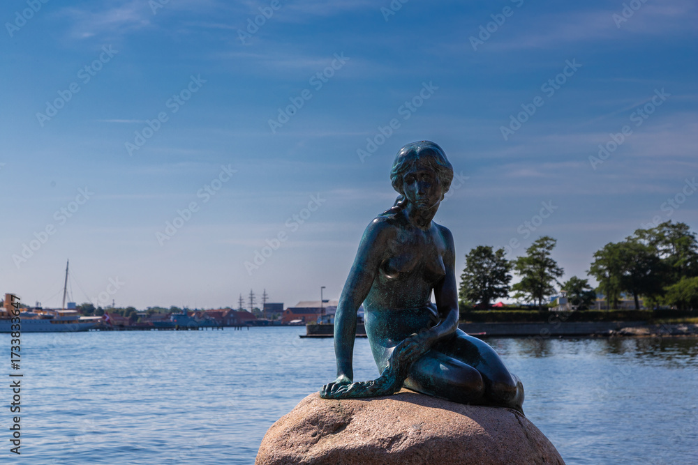 Naklejka premium Copenhagen, Denmark 07-20-2025 The bronze statue of the Little Mermaid sitting on a rock by the waterside at the Langelinie promenade in Copenhagen. The small and unimposing statue is an icon in town.