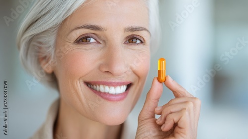An elegant mature woman, exudes vitality and a healthy glow, holding up a single vitamin capsule, symbolizing the positive effects of supplements and wellness.