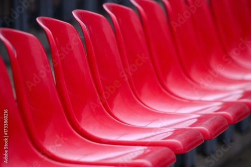 Abstract close-up of glossy red stadium seats forming a rhythmic curve. Selective focus and repetition create a clean graphic pattern. Empty grandstand, no people.