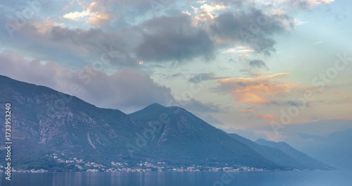 Moonset at calm pre-dawn over Kotor Bay, Montenegro, with misty mountains, soft clouds, and pink-blue sky reflecting in still waters before sunrise 