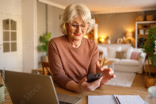 Senior woman with laptop on desk using mobile phone to pay household bills online while sitting at home