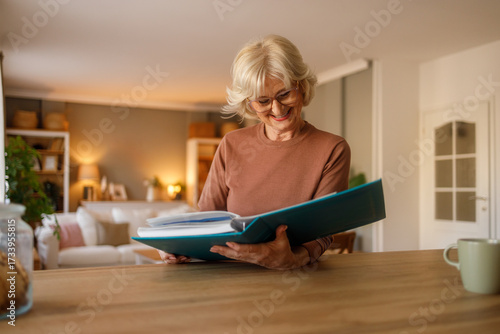 Smiling senior woman looking old photo album and reminiscing family memories while sitting at table in living room